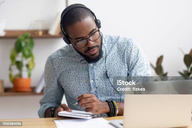 Focused African Businessman In Headphones Writing Notes Watching Webinar Stock Photo - Download Image Now