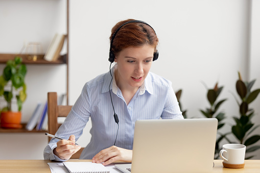 Businesswoman wearing headphones watching video webinar making conference online call writing notes talking on skype, focused woman study online looking at laptop listening translating lecture course