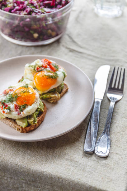 istock Breakfast served of two toasts with avocado, fried eggs with vegetables and herbs on a rustic tablecloth background. View from above. Healthy and nutritious food concept. 1150385336
