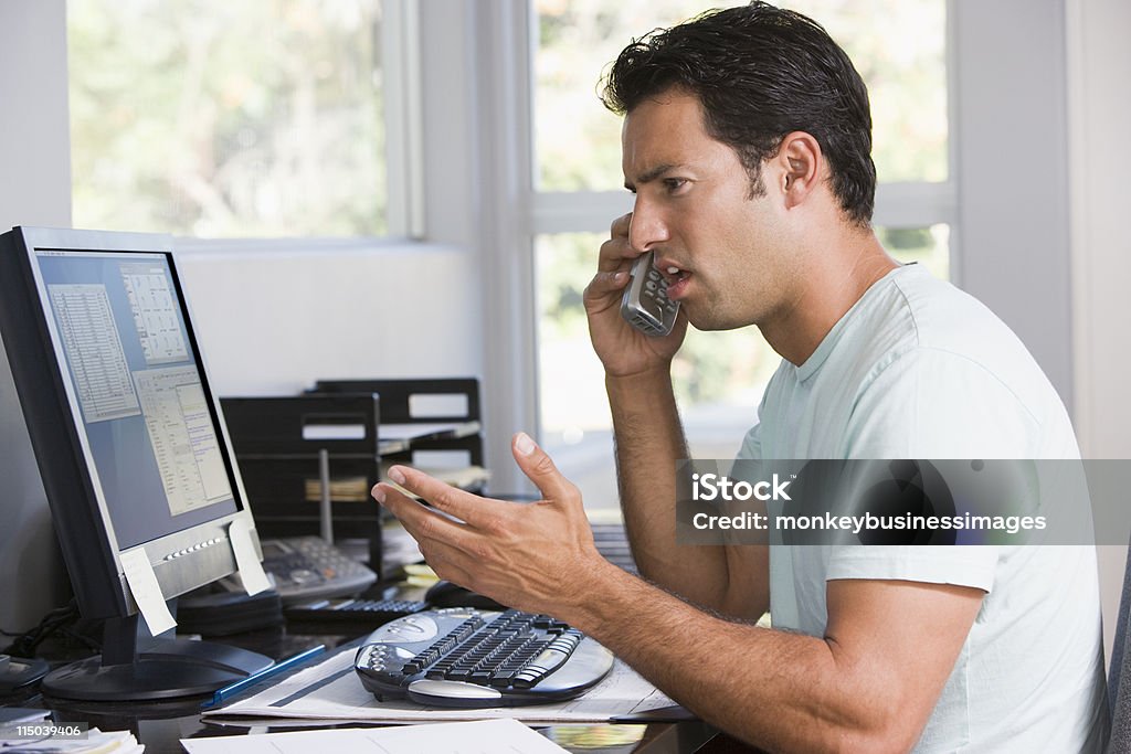 Homem no escritório em casa usando o computador e telefone com o cenho franzido - Foto de stock de Descontente royalty-free