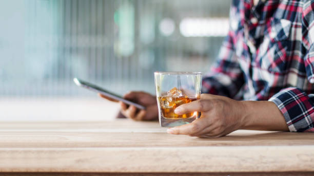 człowiek relaksujący się z bourbon whisky pić napój alkoholowy w ręku i za pomocą telefonu smartphone czat z sieci społecznej w barze w domu, po pracy. - friendship relaxation cocktail nightclub zdjęcia i obrazy z banku zdjęć
