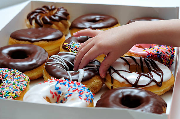 Side view of a young girl's hand reaching in to grab a doughnut from a box of one dozen donuts. 