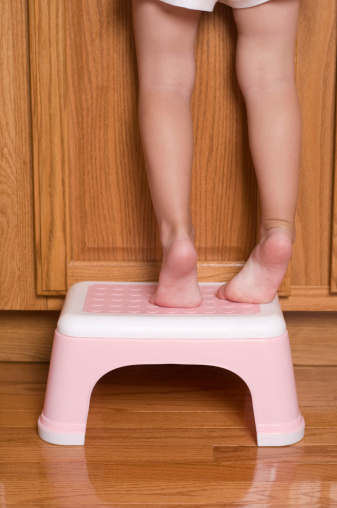 A 3 year old girl's legs and feet tippy toes on a step stool to reach something on a cabinet counter. 