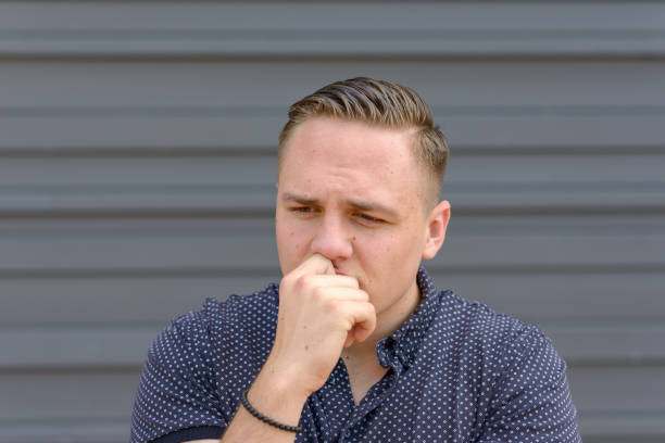 Anxious worried young man frowning as he stands with his hand to his lips and a look of stressed concentration in front of a grey wall