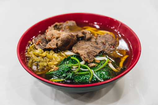 Taiwanese style beef noodle including sliced beef, flat egg noodle, sprouts, morning glory and soup in red bowl.