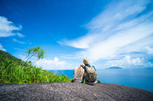 rear view man with backpack, and straw hat  woman with old manual camera, couple sitting relaxing together on granite rock watching the calm indian ocean on sunny vacation day