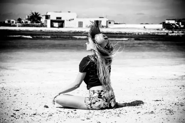 Photo of Black and white romantic and motivational portrait of beautiful blonde girl viewed from back sitting on the sand at the beach with sea and white houses in background - freedom and happiness people