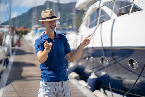 happy smiling 47 years old man with straw hat standing on jetty of luxury marina close to his yacht looking at camera pointing with fingers