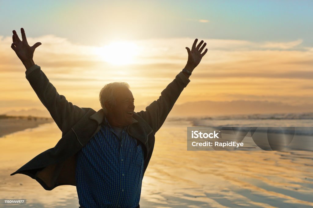 Joyful senior man gestures ecstatically on sunlit beach Almost silhouetted by the setting sun, a senior man stands on a sandy beach gesturing in sheer joy at the beauty of nature around him. Men Stock Photo