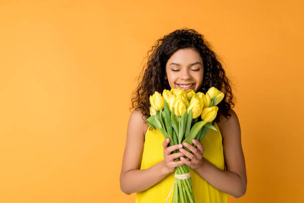 mujer afroamericana rizada y feliz oliendo tulipanes amarillos aislados en naranja - tulip bouquet fotografías e imágenes de stock