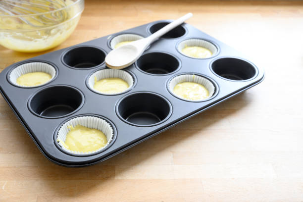 istock muffin tin, half filled with paper cups and dough for cupcake baking on a wooden kitchen table, copy space 1150455133