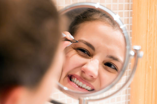 beautiful woman with tweezers is plucking eyebrows while looking into the mirror in bathroom. beauty skincare and wellness morning concept - plucking an instrument imagens e fotografias de stock