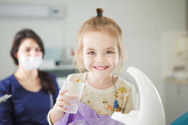 Little child happy after painless teeth polishing procedure Little child happy after painless teeth polishing procedure stands near sink in dentist office with plastic cup with water and female doctor sits behind in facial mask. pediatric dentistry stock pictures, royalty-free photos & images