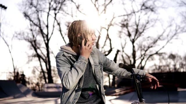 Portrait of happy handsome teenage boy with dreadlocks, using mobile phone while riding a BMX bike, smiling, messaging friends via social networks. People, lifestyle and communication concept