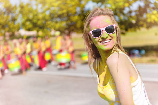 Cheerful young woman enjoying drumming performance on color festival and smiling at camera. Drummer musicians in the background.