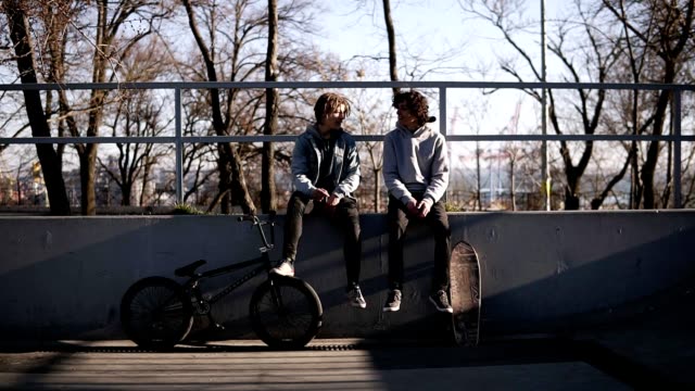 Front view of two friends skateboarder and bmx rider are sitting together on high parapet in the city skate park. Friends are talking, discussing. Sunny day. Active leisure time, sport concept
