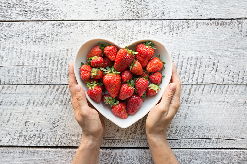 Ripe organic strawberry in plate as heart in female hand on white wooden background. View from above.