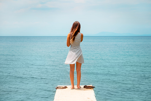 Rear view of teenage girl with long hair standing on a pier, wearing sleeveless dress