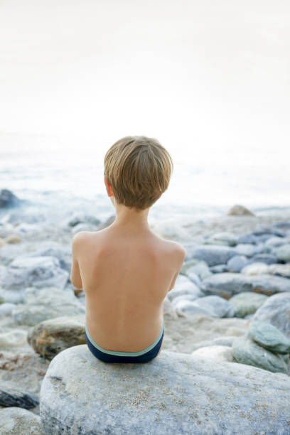 Rear view of a boy in swimming trunks sitting on a rock at the coastline, watching the open sea