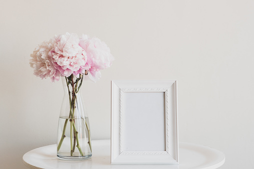 Close up of pale pink peonies in glass vase with blank rectangular picture frame on white table - matte filter effect and selective focus