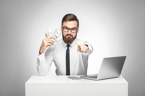 You can earn money! Portrait of handsome satisfied bearded young boss in white shirt and black tie sitting in office, pointing to you finger and holding fan of money,looking at camera, indoor isolated