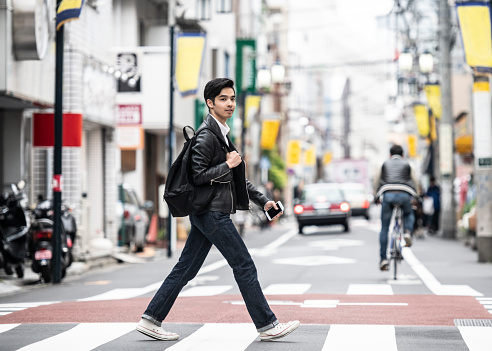 Young Asian man walking across the road looking away and holding phone