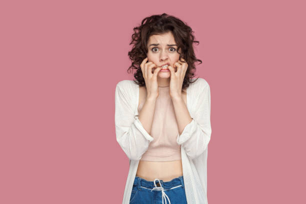 Portrait of nervous worried brunette young woman with curly hairstyle in casual style standing, confused face, bitting her nails and looking at camera. Portrait of nervous worried brunette young woman with curly hairstyle in casual style standing, confused face, bitting her nails and looking at camera. indoor studio shot isolated on pink background. nail biting stock pictures, royalty-free photos & images