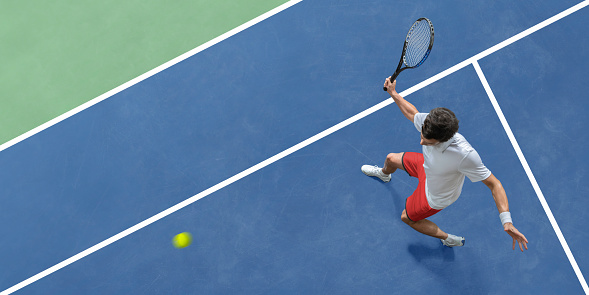 An overhead view of a male tennis player dress in white shirt and red shorts, holding up a racket in mid action, about to strike the ball in a volley during a tennis match. The player is playing on a blue/green tennis court appearing as abstract shapes from above.