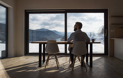 A rear view of mature man sitting at the table in new home, using tablet.