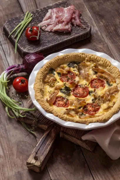 Kish open pie with tomatoes, bacon, spinach in white baking dish on wooden background