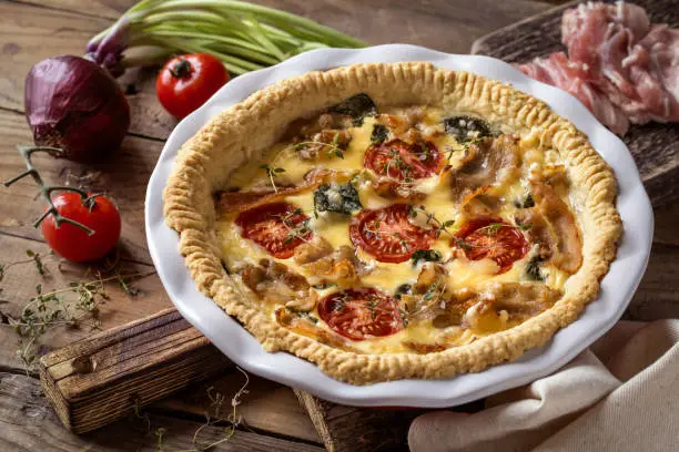 Kish open pie with tomatoes, bacon, spinach in white baking dish on wooden background