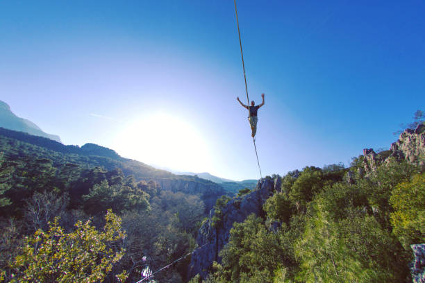 A man is walking along a stretched sling. Highline in the mountains. Man catches balance. Performance of a tightrope walker in nature. Highliner on the background of valley. A man is walking along a stretched sling. Highline in the mountains. Man catches balance. Performance of a tightrope walker in nature. Highliner on the background of valley. highlining stock pictures, royalty-free photos & images