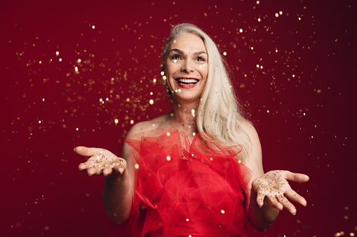 Excited senior woman throwing glitters in studio. Caucasian senior woman playing with golden star glitters over red background.