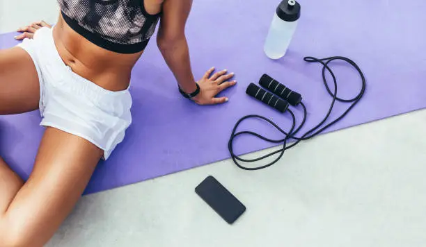 Cropped shot of a woman in fitness wear relaxing after workout sitting on a yoga mat with mobile phone, water bottle and skipping rope beside her.
