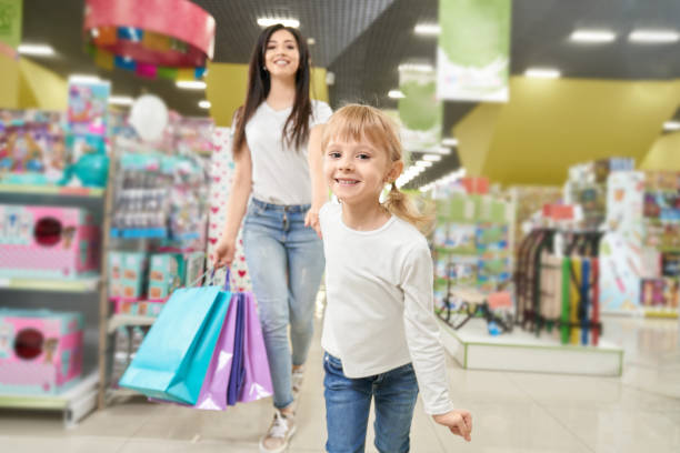 front view of happy girl looking at camera while keeping hand of mother and running forward in big toy store. sming woman following little daughter and laughing. concept of shopping. - boutique shopping retail mother imagens e fotografias de stock