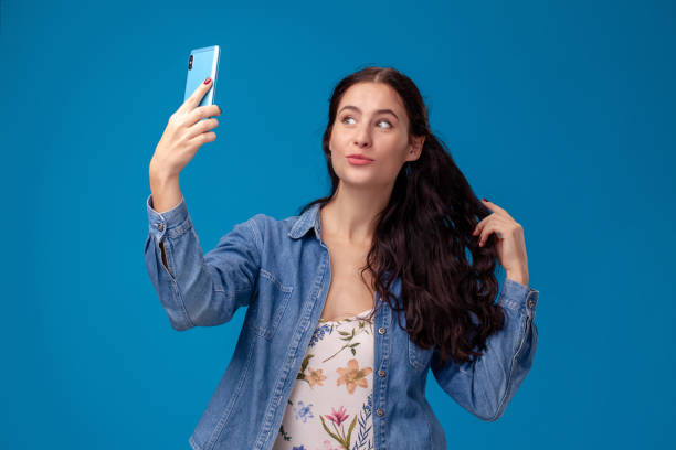 istock Young brunette woman is posing with a smartphone standing on blue background. 1150510635