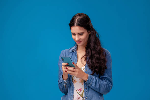 istock Young brunette woman is posing with a smartphone standing on blue background. 1150510649