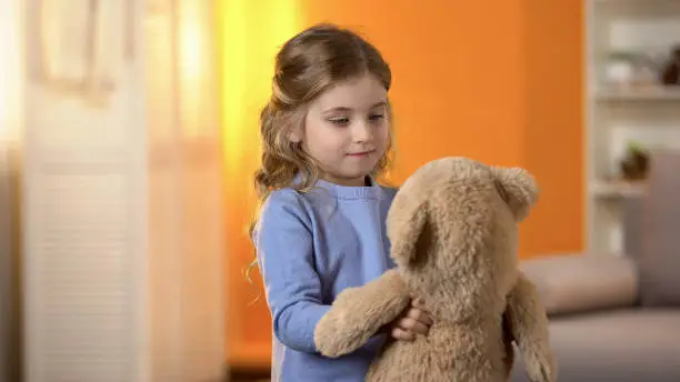 Photo of Happy blond curly-haired little girl playing with favorite teddy bear, childhood