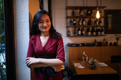 Attractive Chinese woman standing in cafe with crossed arms, leaning on wall, looking at camera, smiling