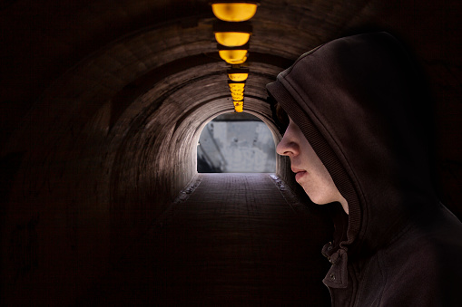 Face of a boy with hood standing alone in a dark underground passage with yellow light bulbs at the roof and light at the end of a tunnel.