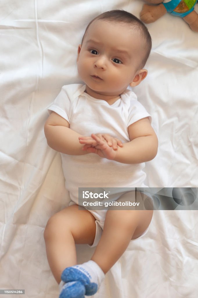6-8 month old baby boy lying playfully in bed 6-8-month-old baby boy lying playfully in bed. Charming 6-7 month little baby in white bodysuit. Baby boy in white bedding. Free space 0-11 Months Stock Photo