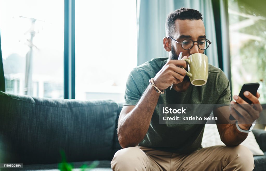 What's the vibe on the newsfeed Shot of a young man using a smartphone and having coffee on the sofa at home Men Stock Photo