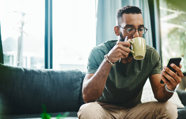 What's the vibe on the newsfeed Shot of a young man using a smartphone and having coffee on the sofa at home coffee break photos stock pictures, royalty-free photos & images