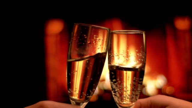 Closeup shoot of two hands of couple in love clinking glasses full of champagne celebrating a night date with cozy warm fireplace on the background indoors