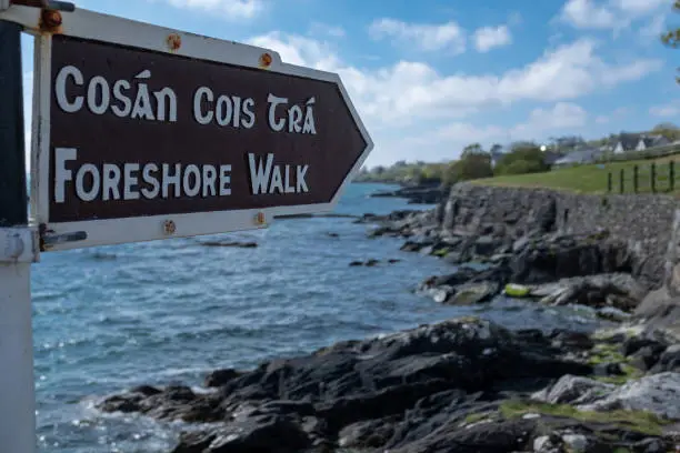 A foreshore walk sign in English and Irish, pointing to the walk along the jagged cliff with the Atlantic ocean crashing on the rocks against a bright blue sky