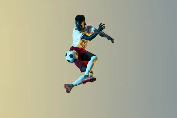 Young african-american male football or soccer player in sportwear and boots kicking ball for the goal in jump in neon light on gradient background. Concept of healthy lifestyle, professional sport.