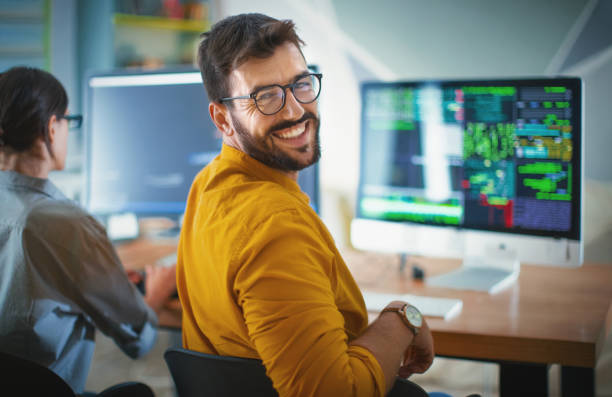 Successful IT developer. Closeup over the shoulder view of a cheerful mid 20's software developer at the office smiling to the camera. His female coworker is next him, still focused and working. looking over shoulder stock pictures, royalty-free photos & images