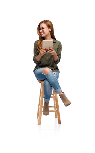 A portrait of a young woman sitting on a wooden stool and holding a tablet while looking over at a copy space.