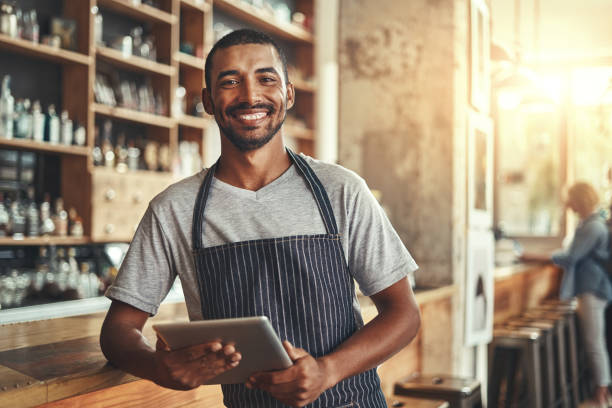 Smiling male entrepreneur in his coffee shop holding digital tab Startup successful male entrepreneur standing in his coffee shop at the counter holding digital tablet in hand cafe tablet stock pictures, royalty-free photos & images