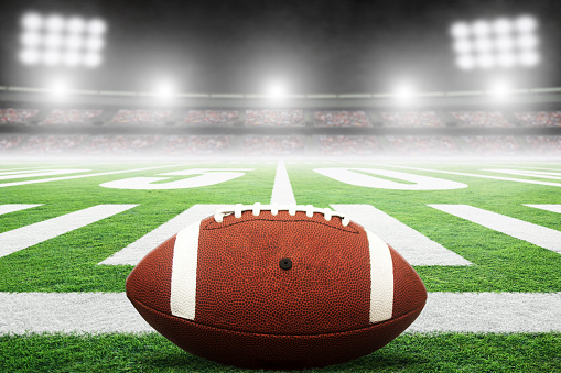 Close up of American football on stadium field with yard line markings and spotlight with blurred background and copy space. Fictitious stadium created in Photoshop.